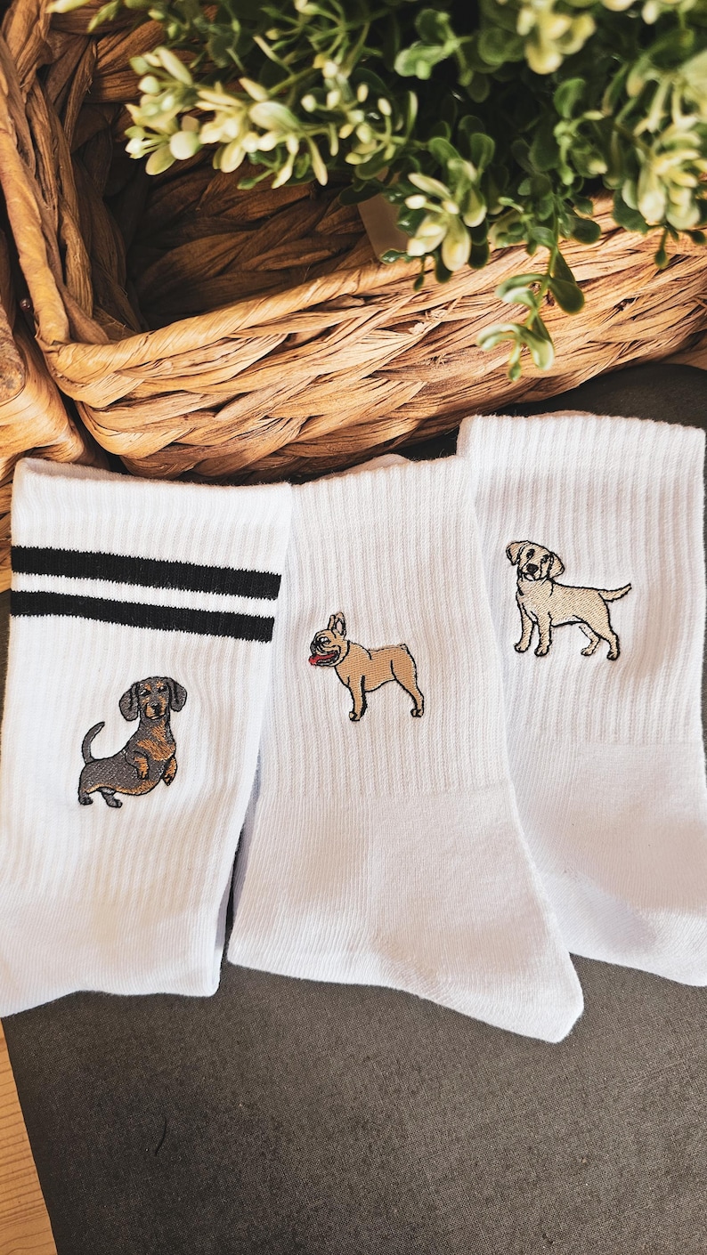 May include: Three pairs of white crew socks with embroidered dog breeds. One pair features a dachshund, another a French bulldog, and the last a Labrador retriever.