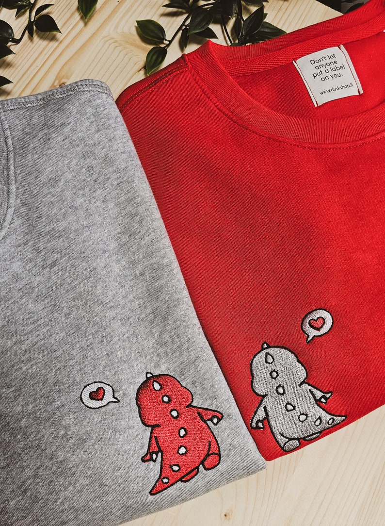 May include: Two crewneck sweatshirts, one gray and one red, are displayed. Each features an embroidered dinosaur design with a heart-shaped speech bubble. The red sweatshirt has a tag that reads, "Don't let anyone put a label on you."