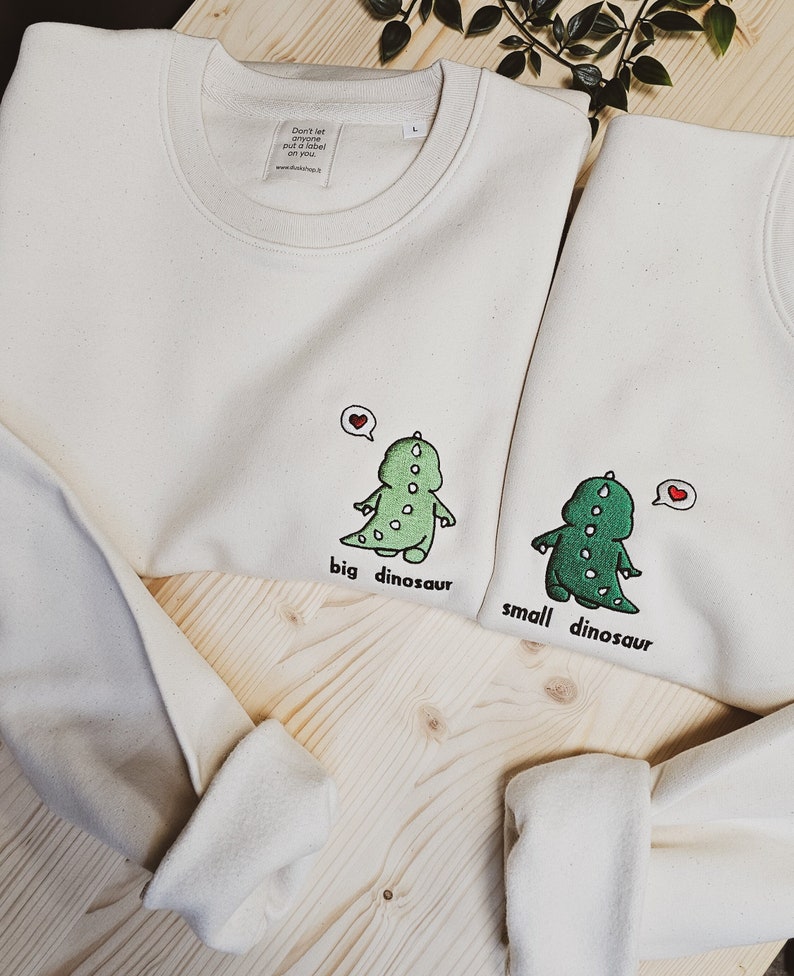 May include: Two off-white sweatshirts with crew necks. Each sweatshirt features a green dinosaur graphic with the words "big dinosaur" and "small dinosaur" below. A red heart is above each dinosaur. The tag reads "Don't let anyone put a label on you."