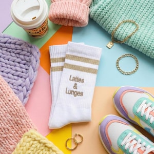 May include: A flat lay arrangement with white socks featuring gold stripes and the text "Lattes & Lunges", alongside pastel-colored knitted items, a coffee cup, and colourful trainers. Gold jewellery and a pastel palette create a coordinated look.