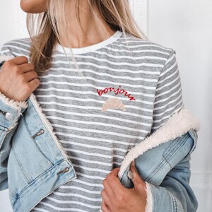 Puede incluir: Una camiseta a rayas grises y blancas con un "bonjour" bordado en rojo y un gr&aacute;fico de croissant.