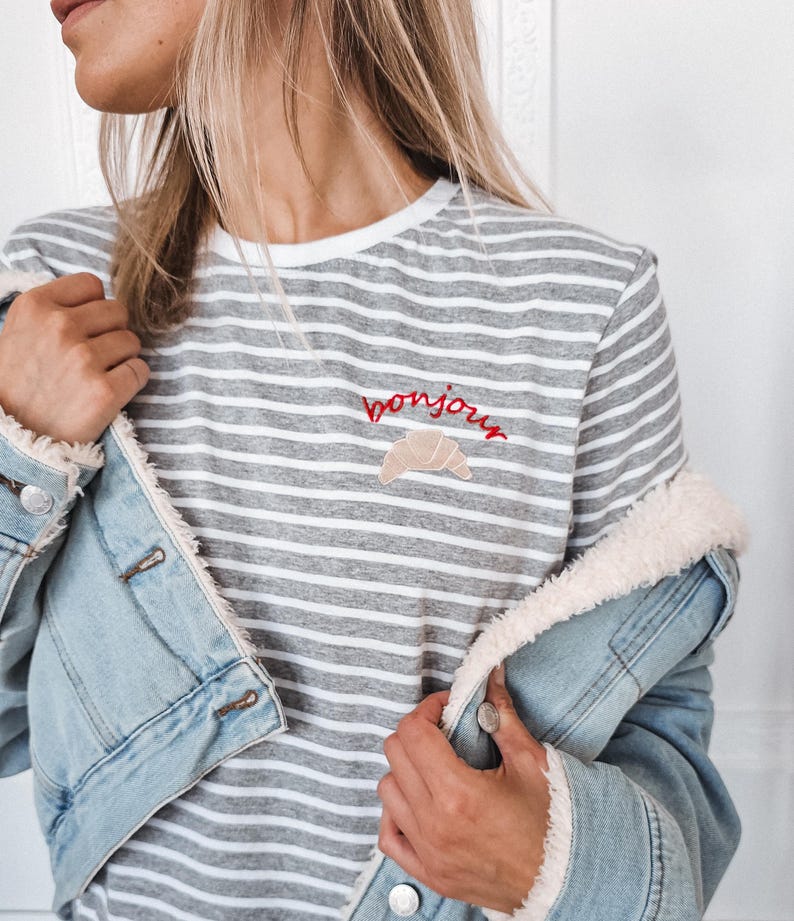 Puede incluir: Una camiseta a rayas grises y blancas con un "bonjour" bordado en rojo y un gr&aacute;fico de croissant.