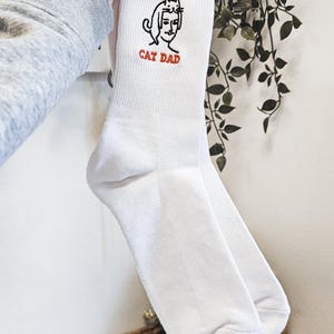 Peut inclure: Chaussettes blanches avec une illustration brod&eacute;e en noir et blanc d'une personne avec un chat sur la t&ecirc;te. Le texte "CAT DAD" est brod&eacute; en rouge sous l'illustration.