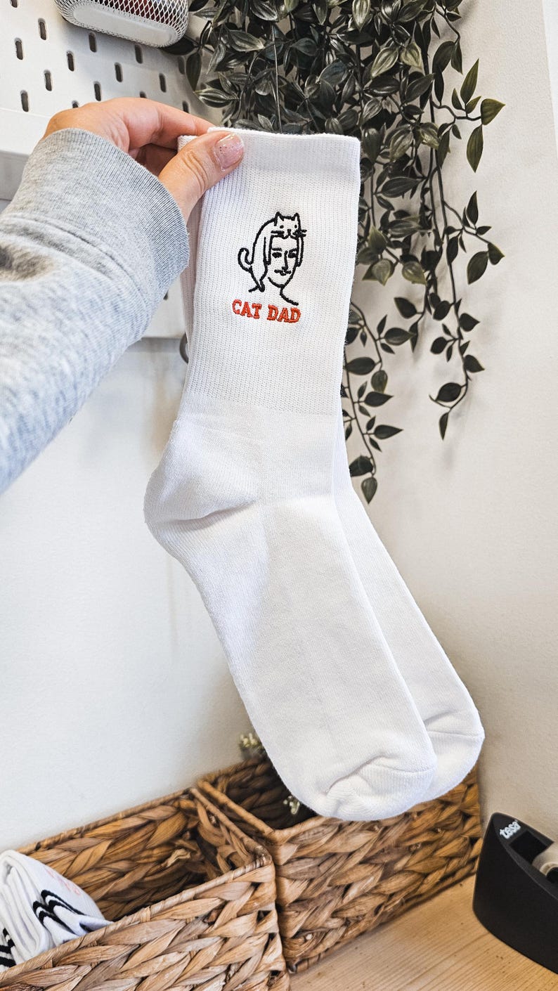 Peut inclure: Chaussettes blanches avec une illustration brod&eacute;e en noir et blanc d'une personne avec un chat sur la t&ecirc;te. Le texte "CAT DAD" est brod&eacute; en rouge sous l'illustration.