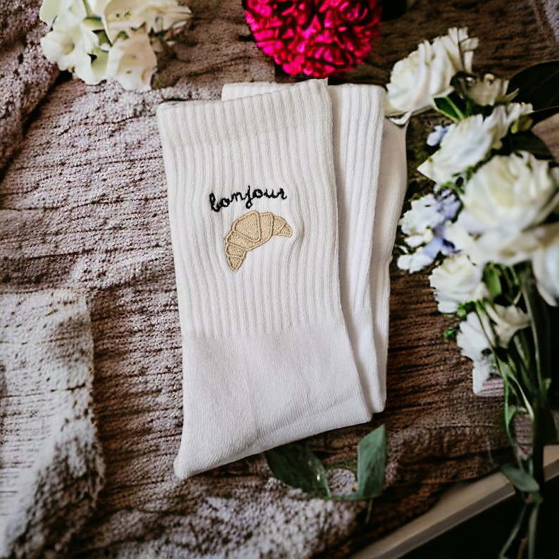 Puede incluir: Calcetines de algod&oacute;n blanco con un croissant bordado en marr&oacute;n y la palabra "bonjour" en negro.