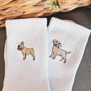 May include: A pair of white socks with embroidered images of a French Bulldog and a Labrador Retriever. The socks are on a grey surface.