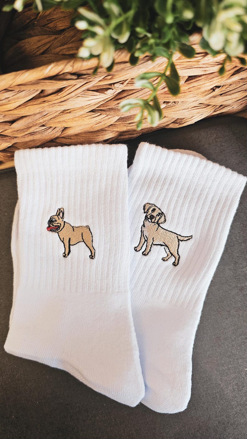 May include: A pair of white socks with embroidered images of a French Bulldog and a Labrador Retriever. The socks are on a grey surface.
