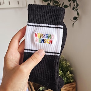 Könnte beinhalten: Schwarze Socken mit einem weißen Band mit dem gestickten Text "HERZENS MENSCH" in Regenbogenfarben. Die Socken haben eine gerippte Textur und ein klassisches Design.