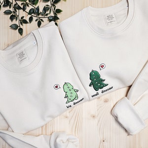 May include: Two off-white sweatshirts with dinosaur graphics. One says "big dinosaur" and the other says "small dinosaur". Each dinosaur has a heart above its head. The sweatshirts have a crew neck and long sleeves.