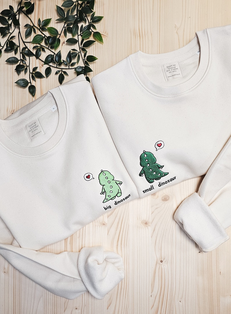 May include: Two off-white sweatshirts with dinosaur graphics. One says "big dinosaur" and the other says "small dinosaur". Each dinosaur has a heart above its head. The sweatshirts have a crew neck and long sleeves.