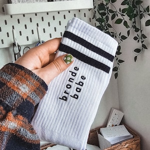 Pode incluir: Meias brancas com listras pretas e o texto "bronde babe" bordado nelas. As meias estão sendo seguradas por uma mão com esmalte de unha verde e branco.