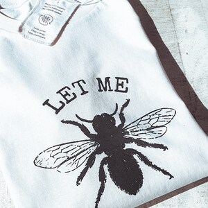 May include: White t-shirt with a black graphic of a bee and the words "LET ME" printed above it. The shirt has a tag attached. The shirt is made from organic textile waste and raw organic cotton.