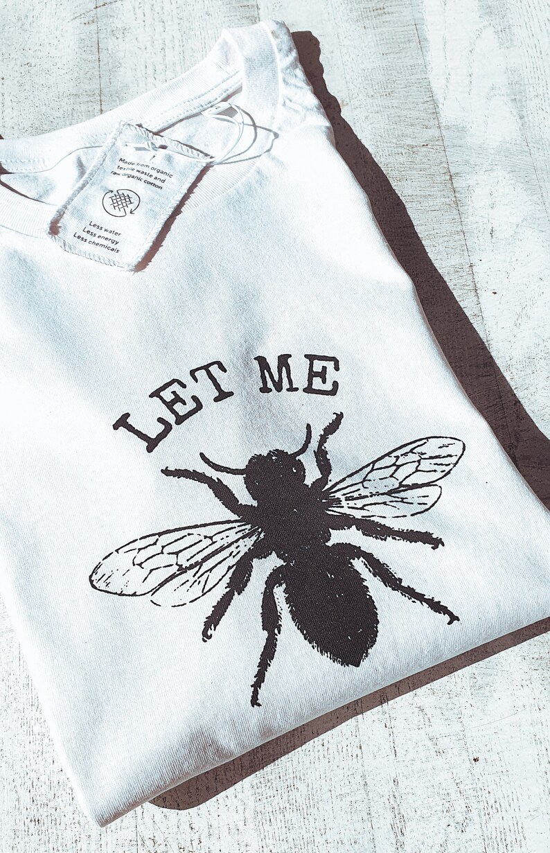May include: White t-shirt with a black graphic of a bee and the words "LET ME" printed above it. The shirt has a tag attached. The shirt is made from organic textile waste and raw organic cotton.