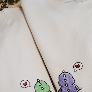May include: Two off-white sweatshirts with embroidered dinosaur designs. One dinosaur is green, the other purple, both with white spots. Each dinosaur has a speech bubble with a red heart.