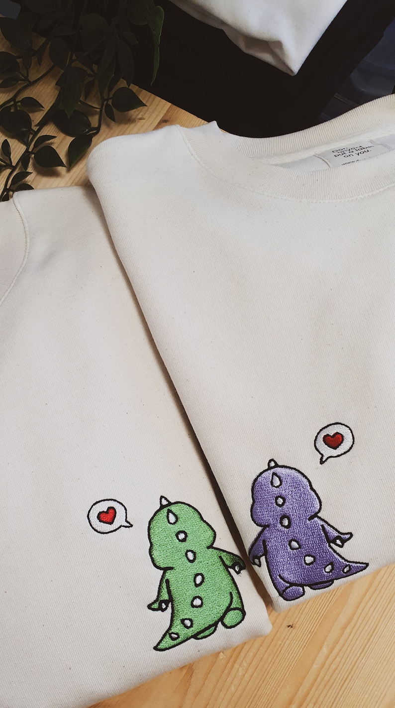 May include: Two off-white sweatshirts with embroidered dinosaur designs. One dinosaur is green, the other purple, both with white spots. Each dinosaur has a speech bubble with a red heart.
