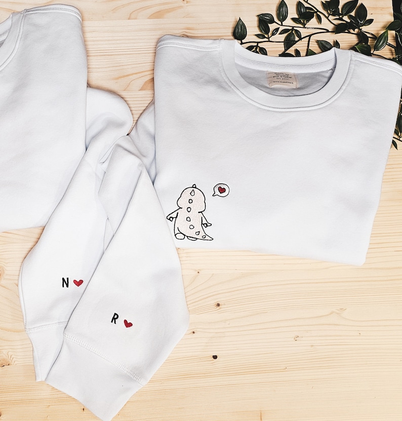 May include: White sweatshirts with embroidered designs. One sweatshirt features a cartoon dinosaur with a heart-shaped thought bubble. Another has the letters "N" and "R" with heart symbols. The sweatshirts are on a wooden surface.