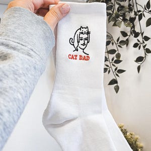 Peut inclure: Chaussettes blanches avec une illustration en noir et blanc d'un chat sur la t&ecirc;te d'une personne. Le texte "CAT DAD" est brod&eacute; en rouge sous l'illustration.