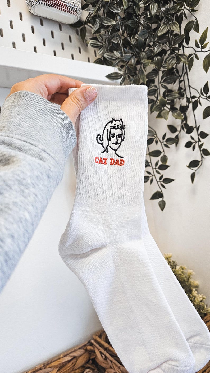 Peut inclure: Chaussettes blanches avec une illustration en noir et blanc d'un chat sur la t&ecirc;te d'une personne. Le texte "CAT DAD" est brod&eacute; en rouge sous l'illustration.