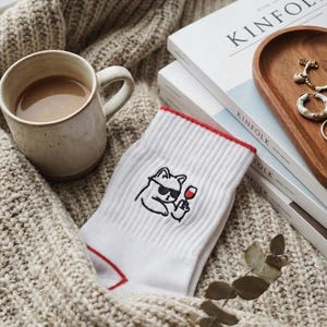 May include: White socks with a red trim and a cartoon cat wearing sunglasses and holding a wine glass. The socks are next to a mug of coffee, books, and jewelry on a cozy, knitted blanket.
