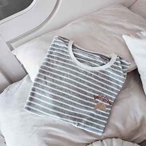 Puede incluir: Una camiseta a rayas grises y blancas con un croissant bordado en marr&oacute;n y el texto "bonjour" en hilo rojo. La camiseta est&aacute; doblada y colocada en una cama blanca con una almohada blanca y una almohada redonda beige.