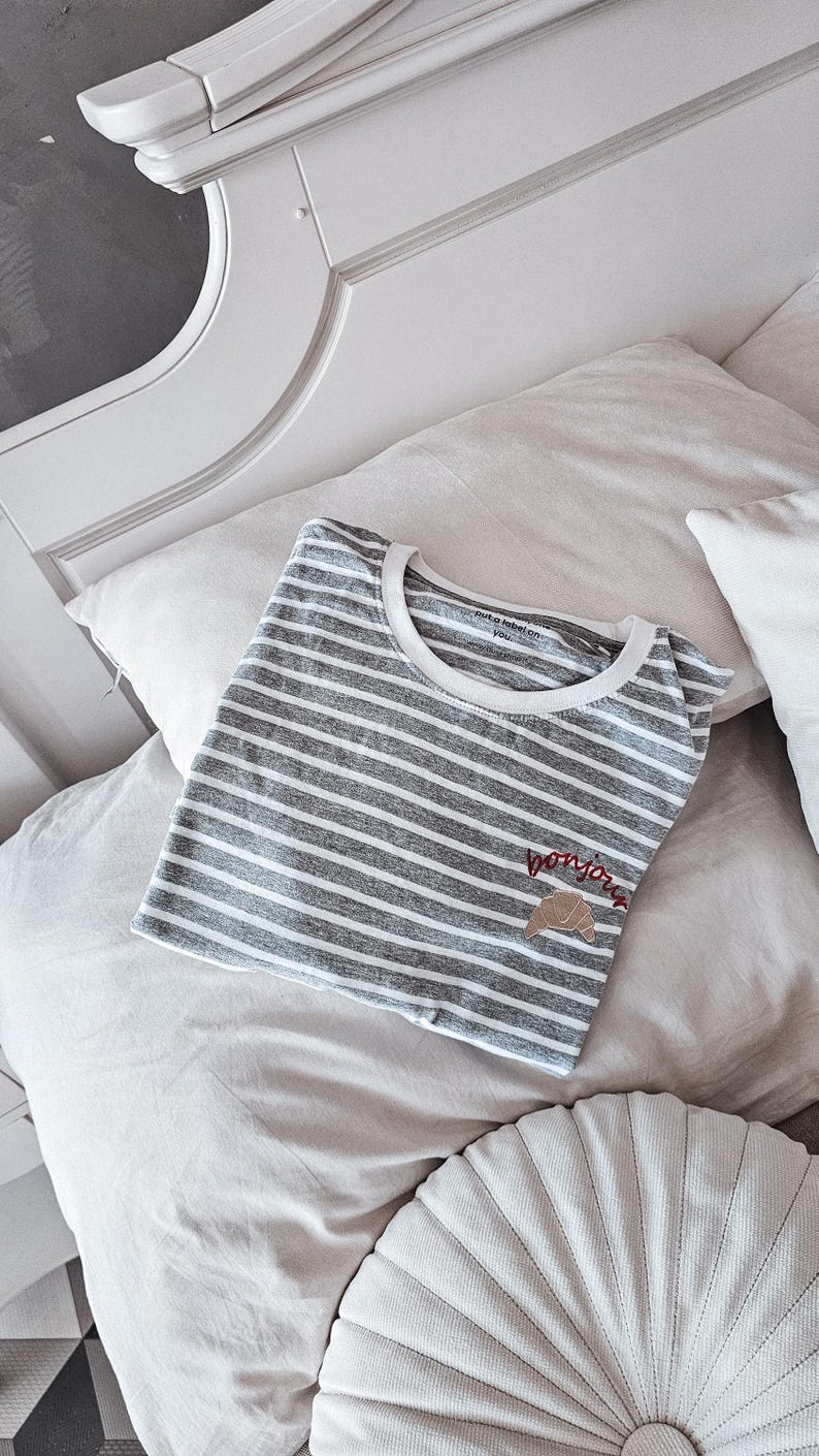 Puede incluir: Una camiseta a rayas grises y blancas con un croissant bordado en marr&oacute;n y el texto "bonjour" en hilo rojo. La camiseta est&aacute; doblada y colocada en una cama blanca con una almohada blanca y una almohada redonda beige.