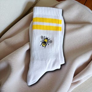 Puede incluir: Calcetines blancos con rayas amarillas y un bordado de abeja. Los calcetines tienen una textura acanalada y un puño. La abeja bordada es negra y amarilla. Los calcetines están sobre una tela de color neutro.