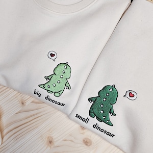 May include: Two off-white sweatshirts with embroidered green dinosaurs and the phrases "big dinosaur" and "small dinosaur." Each dinosaur has a heart above its head. The sweatshirts are on a light wood surface.