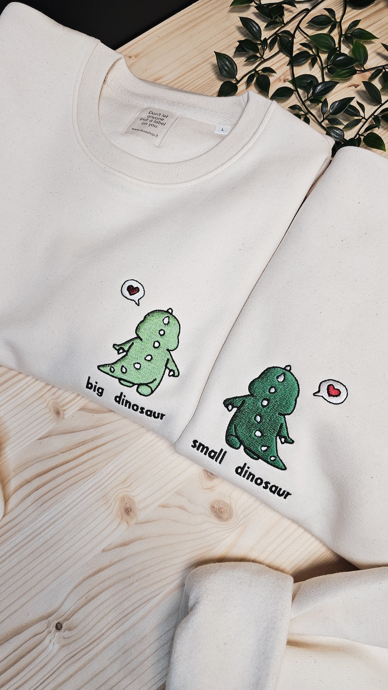 May include: Two off-white sweatshirts with embroidered green dinosaurs and the phrases "big dinosaur" and "small dinosaur." Each dinosaur has a heart above its head. The sweatshirts are on a light wood surface.