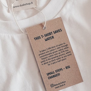May include: A brown paper tag with white text that reads "This T-shirt saves water." The tag explains that it takes 2,700 litres of water to produce one cotton t-shirt through conventionally grown cotton, while organic cotton uses up 91% less water in the growing process. The tag also says "Small steps - big changes!" and includes the social media handle @duskclothes and website www.dusk.lt.