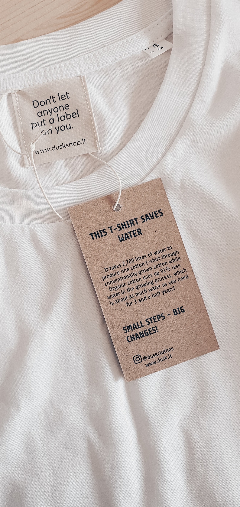 May include: A brown paper tag with white text that reads "This T-shirt saves water." The tag explains that it takes 2,700 litres of water to produce one cotton t-shirt through conventionally grown cotton, while organic cotton uses up 91% less water in the growing process. The tag also says "Small steps - big changes!" and includes the social media handle @duskclothes and website www.dusk.lt.