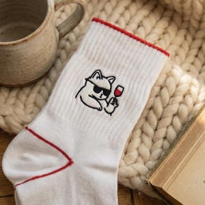 May include: White socks with a red trim and a cartoon cat wearing sunglasses and holding a wine glass. The socks are on a cream-colored knitted blanket, with a ceramic mug and a book in the background.