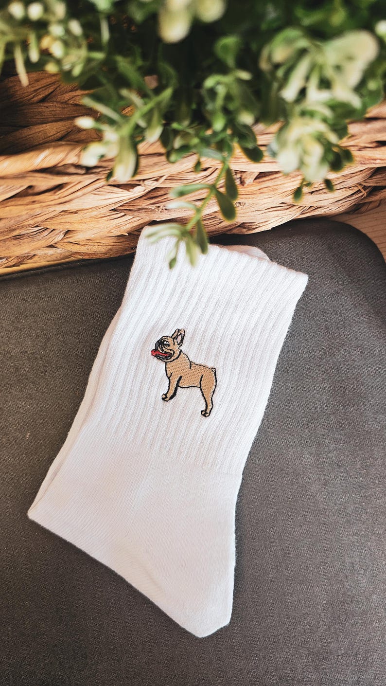 May include: White cotton socks with an embroidered image of a French bulldog. The dog is brown with a black nose and a red tongue.