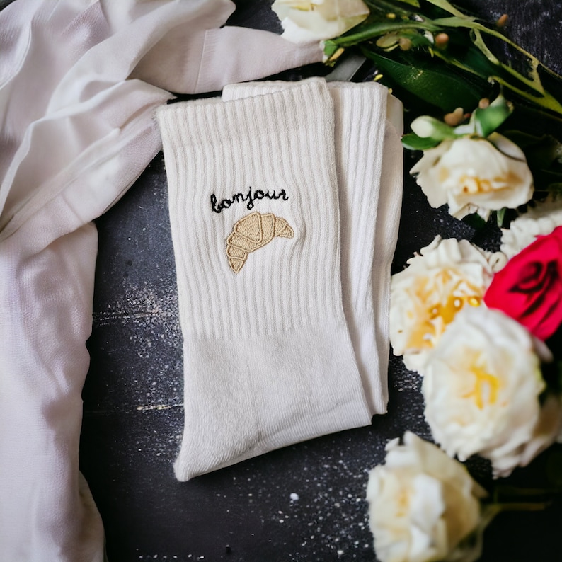 Puede incluir: Calcetines blancos de algod&oacute;n con un croissant bordado en negro y la palabra "bonjour" en una de las calcetines.