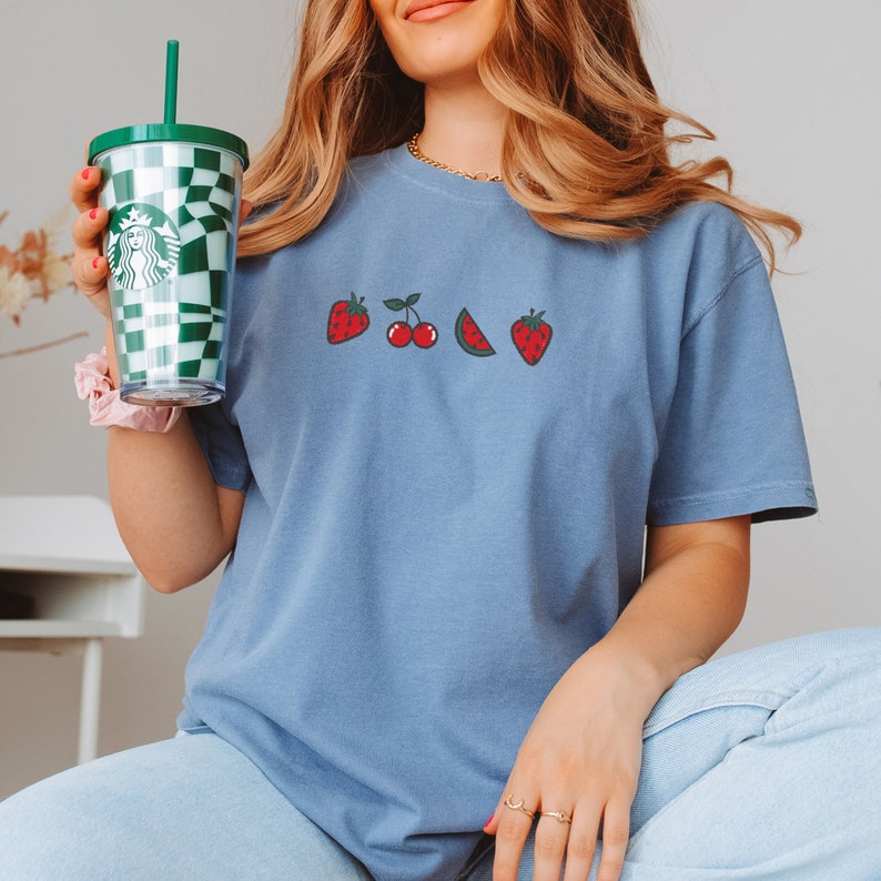 May include: A light blue t-shirt with a graphic of strawberries, cherries, and a watermelon. The shirt is being held by a person who is also holding a green and white checkered cup with a green straw.