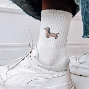 May include: White trainers with a white sock featuring an embroidered dachshund. The sock is worn with blue denim jeans.