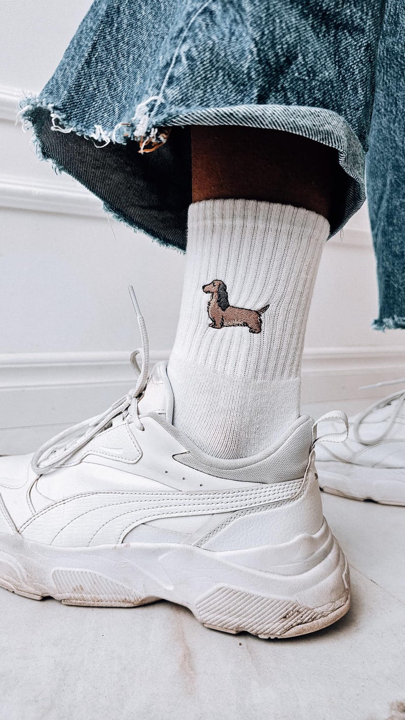 May include: White trainers with a white sock featuring an embroidered dachshund. The sock is worn with blue denim jeans.