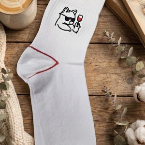 May include: White crew sock with a red trim and a cartoon cat wearing sunglasses and holding a glass of wine. The sock is on a wooden surface with a cup of coffee and a book.