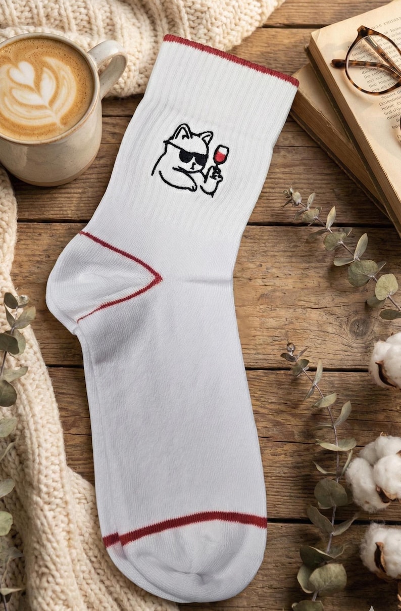 May include: White crew sock with a red trim and a cartoon cat wearing sunglasses and holding a glass of wine. The sock is on a wooden surface with a cup of coffee and a book.