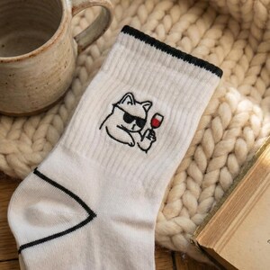 May include: White socks with a black trim and a black cat graphic wearing sunglasses and holding a glass of red wine. The socks are on a wooden surface next to a mug and a book.
