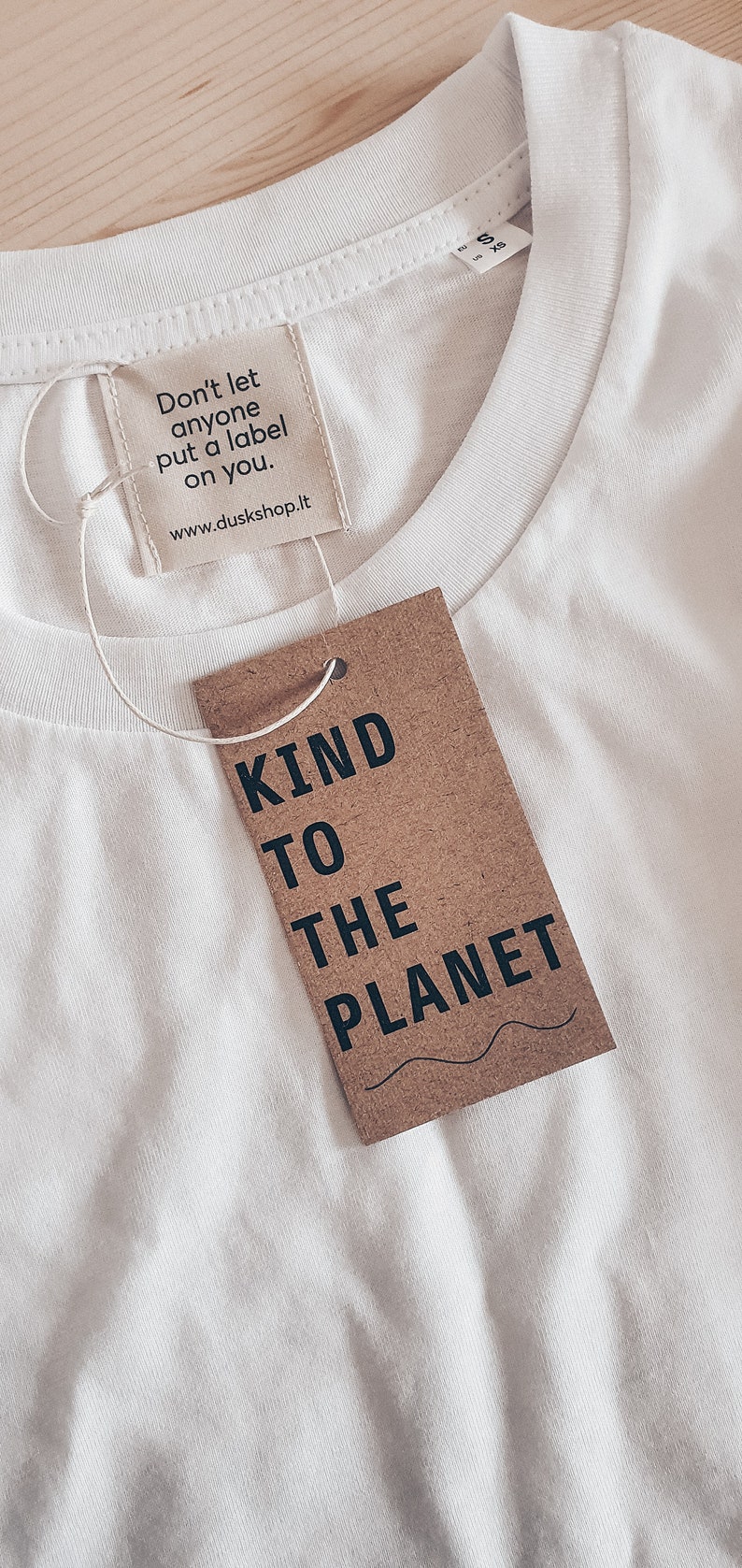 May include: White t-shirt with a brown tag that says "Kind to the Planet". The tag is attached to the shirt with a white string. The shirt also has a smaller tag that says "Don't let anyone put a label on you. www.duskshop.it"