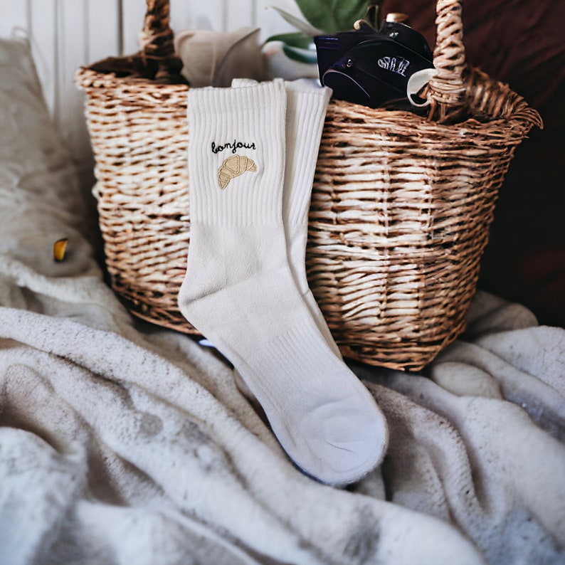 Puede incluir: Un par de calcetines blancos con un croissant y la palabra "bonjour" bordada en ellos. Los calcetines est&aacute;n sobre una manta blanca junto a una cesta de mimbre.