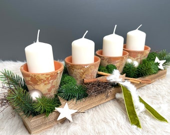 May include: A rectangular wooden tray holding four terracotta pots, each containing a white pillar candle. The arrangement is decorated with green pine branches, moss, cinnamon sticks, white star ornaments, and a green velvet ribbon.