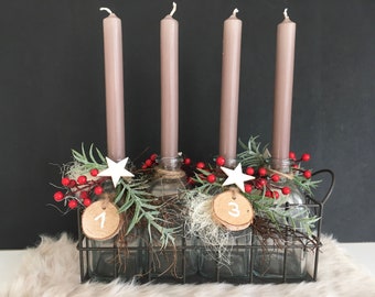 May include: Four tall taupe taper candles placed in clear glass bottles inside a dark metal wire basket. The bottles are decorated with red berries, green foliage, white wooden stars, and round wooden tags labeled with the numbers 1 and 3.