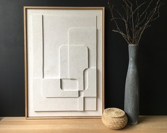 May include: A rectangular wall art piece featuring a white textured surface with raised geometric shapes in a light wood frame. Beside it sits a tall, slender gray vase containing bare branches and a small, round, woven tan basket with a lid.