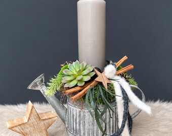 May include: A galvanized metal watering can centerpiece featuring a tall beige pillar candle, a green succulent, cinnamon sticks, pine needles, moss, and white and dark gray felt strips, accompanied by a separate wooden star ornament.