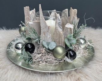 May include: A decorative centerpiece featuring a clear glass candle holder surrounded by vertical pieces of light-colored bark, accented with silver and black spherical ornaments, green eucalyptus leaves, and wispy fibers on a round silver tray.