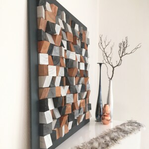 May include: A geometric wall art piece made of wood blocks in shades of gray, brown, and white. The blocks are arranged in a random pattern to create a modern and abstract design. The art piece is mounted on a black background and is displayed on a white shelf.