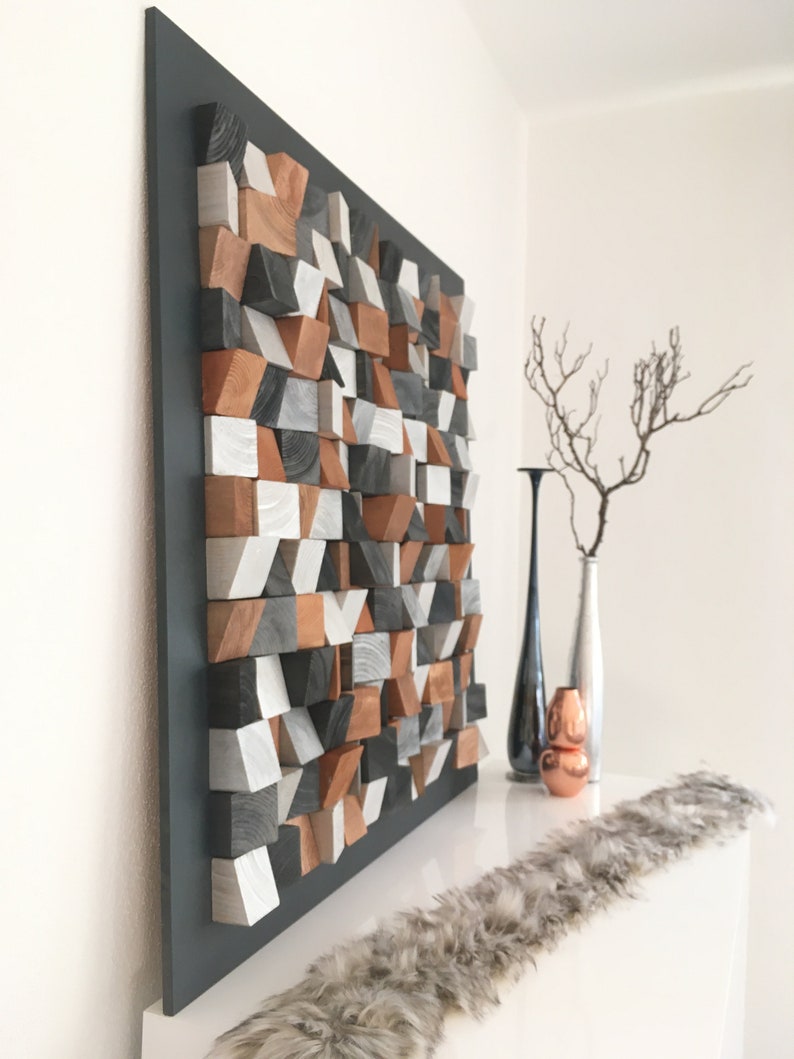 May include: A geometric wall art piece made of wood blocks in shades of gray, brown, and white. The blocks are arranged in a random pattern to create a modern and abstract design. The art piece is mounted on a black background and is displayed on a white shelf.