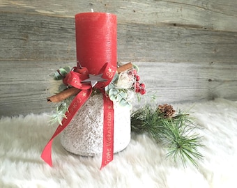 May include: A red pillar candle sits atop a textured white base, decorated with a red ribbon printed with the text Frohe Weihnachten, a wooden star, cinnamon sticks, artificial red berries, and green foliage accents.