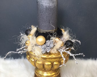 May include: A tall, textured gray pillar candle sits inside a gold-finished urn. The base of the candle is decorated with a cluster of gold and black spheres, silver-toned berry accents, and wispy, light-colored dried botanical elements.